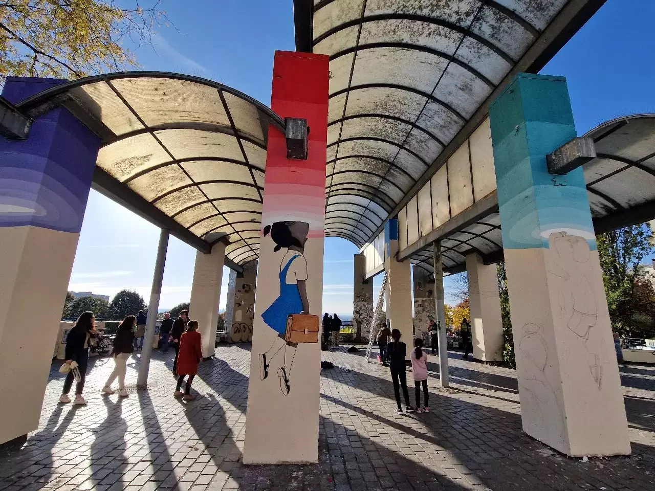 Piliers peints de street art sous un auvent, dont une fillette au cartable, avec des passants au soleil