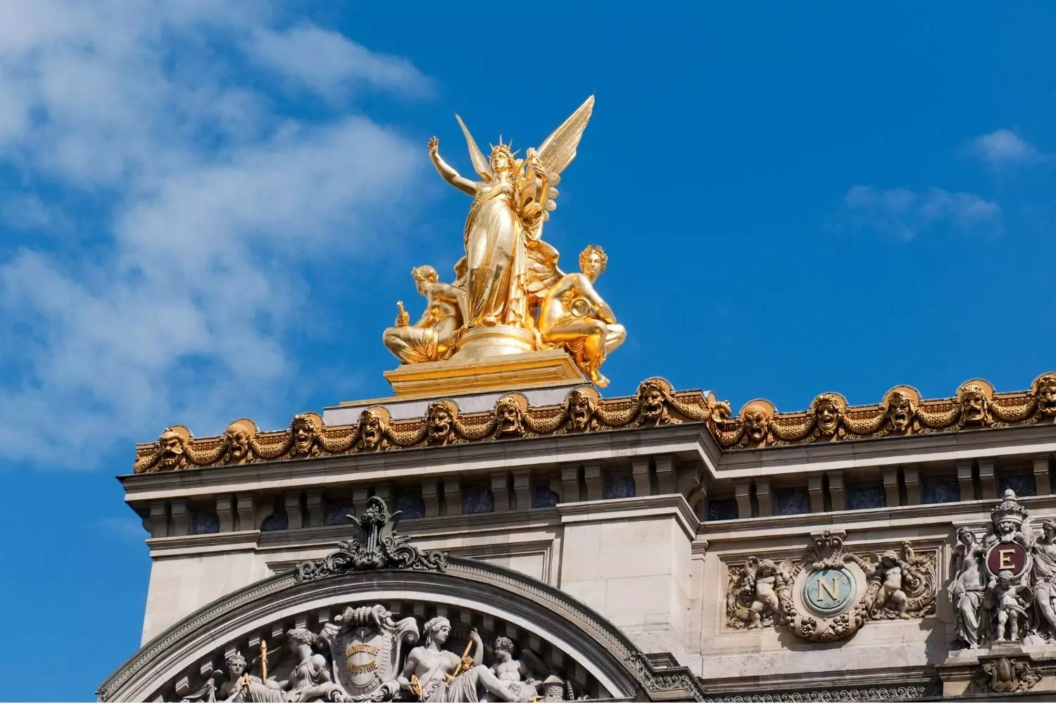 Gilded sculpture group at the top of the Palais Garnier facade, under a blue sky