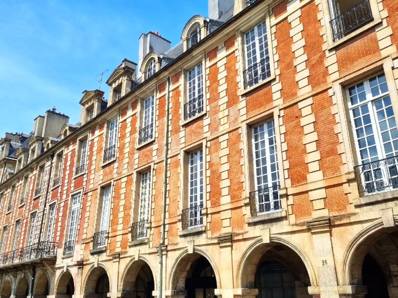 Brick and stone facades and arcades of the Place des Vosges under a blue sky