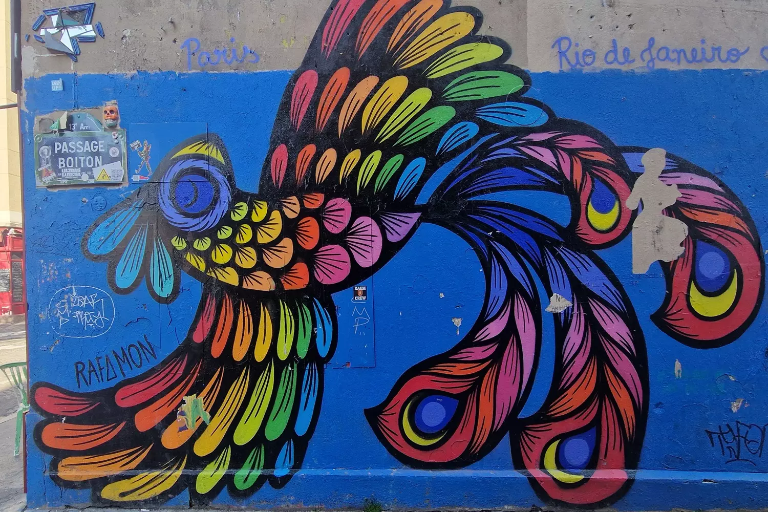 Fresque de street art d'un oiseau aux plumes multicolores sur un mur bleu, passage Boiton